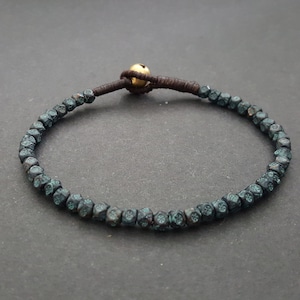 Pode incluir: Um bracelete azul escuro com um cordão castanho e uma conta dourada. As contas são pequenas e têm uma superfície texturizada.
