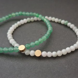 May include: Two bracelets made of green and white beads with gold accents. The bracelets are stacked on top of each other.