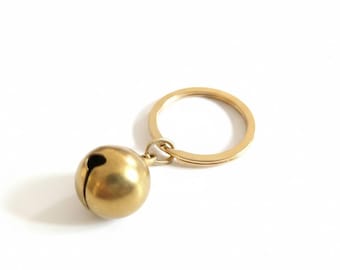 Puede incluir: Un llavero dorado con una pequeña campana redonda con una ranura vertical. La campana está unida a un llavero circular. El llavero está hecho de un material metálico brillante sobre un fondo blanco.
