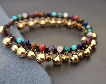 Pode incluir: Um bracelete de cordão castanho com sinos dourados e contas de pedras preciosas coloridas. As contas estão em um padrão de arco-íris, com contas turquesa, azuis, roxas, vermelhas, laranja, amarelas e verdes.