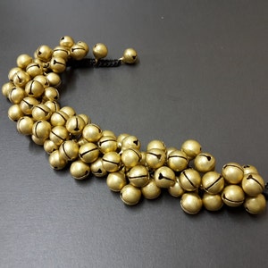 May include: A gold-colored bracelet with a string of small bells attached. The bracelet is made of a black cord and has a simple design.