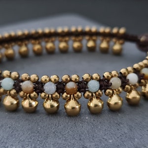 May include: A brown and gold beaded anklet with small gold bells. The anklet is made with brown cord and features a variety of light-colored beads.