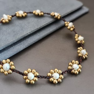 Op de afbeelding: Een bruin koordarmband met gouden en witte bloemvormige kralen. De armband heeft twee kleine gouden belletjes.
