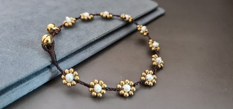 Op de afbeelding: Een bruin koordarmband met gouden en witte bloemvormige kralen. De armband heeft twee kleine gouden belletjes.