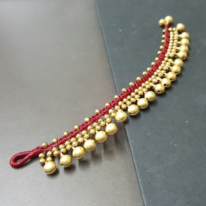 May include: Red and gold ankle bracelet with small bells. The bracelet is made of red string and has gold bells attached to it. The bells are arranged in a cascading pattern.