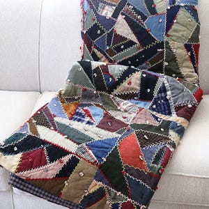 May include: A patchwork quilt with a variety of fabric pieces in shades of blue, red, green, and brown. The quilt is made of triangular and rectangular shapes, stitched together with white thread. The quilt is folded over a white sofa.