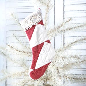 May include: A small, quilted Christmas stocking with a red and white patchwork design. The stocking is decorated with a cream-colored lace trim at the top and hangs from a white tree branch. The stocking is approximately 6 inches long.