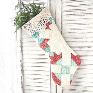 May include: A quilted Christmas stocking featuring a cream-colored base with a geometric pattern in red and teal. The top cuff is adorned with white crocheted lace trim, and the interior is filled with a sprig of green evergreen foliage.
