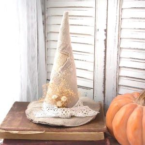 May include: A conical hat crafted from light beige burlap fabric. The hat features a wide brim and is decorated with a white crocheted lace ribbon tied in a bow at the base, accented with a small cluster of dried beige baby's breath flowers.