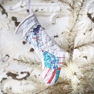 May include: A small, quilted Christmas stocking with a patchwork design. The stocking features a white background with sections of blue, red, and floral patterns. A white lace trim adorns the top edge, and a white ribbon is attached for hanging.