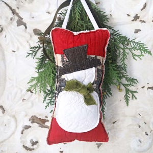 May include: A handmade snowman ornament with a red top and bottom, a black hat, and a white body. The snowman has a green scarf and is hanging from a white ribbon. The background includes evergreen branches.