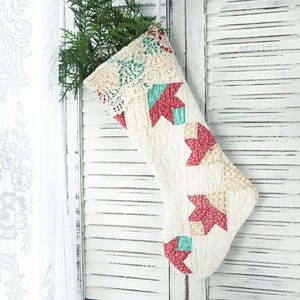 May include: A quilted Christmas stocking featuring a cream-colored base with a repeating geometric pattern in shades of red and teal. The top cuff is adorned with intricate cream lace trim, and green pine branches protrude from the opening.