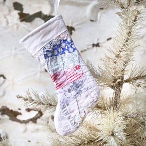 May include: A small, handmade Christmas stocking with a patchwork design. The stocking features various fabric patterns in shades of blue, red, and white, with a white lace trim at the top. A white ribbon is attached for hanging.