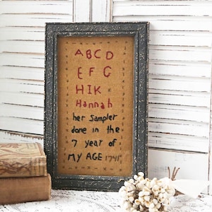 May include: Framed antique sampler with the text "ABCD EFG HIK Hannah her Sampler done in the 7 year of MY AGE 1741." The sampler is in a black frame with a distressed finish. The background is a tan color.