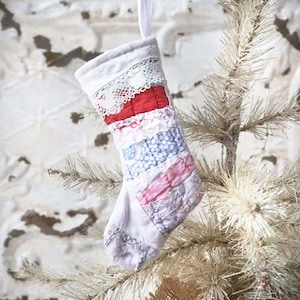 May include: A small, handmade Christmas stocking with a patchwork design. The stocking features a white lace trim at the top, a red band, and various patterned fabric strips in blue, pink, and white. A white ribbon is attached for hanging.
