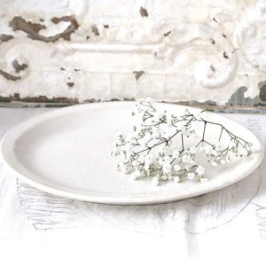May include: A white, oval-shaped ceramic serving platter with a slightly raised rim. Delicate white baby's breath flowers rest on the platter. The background features a distressed white wall and a white linen tablecloth.