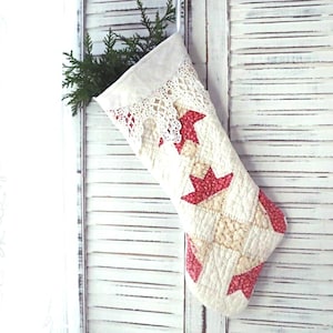 May include: A quilted Christmas stocking featuring a cream-colored fabric base with a repeating red and white star pattern. The top cuff is accented with white lace trim, and the interior contains a small sprig of green evergreen foliage.