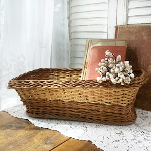 May include: A rectangular woven wicker basket with a light brown finish, featuring a braided rim and a tapered base. The basket is filled with a small cluster of white dried flower buds and two vintage books with worn, orange-toned covers.