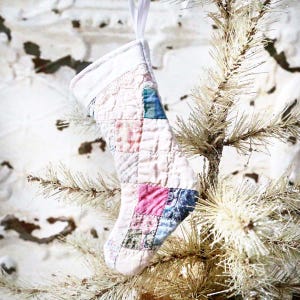 May include: A quilted Christmas stocking with a patchwork design in shades of pink, blue, and green. The stocking is hanging from a white ribbon on a white Christmas tree. The fabric has a textured, vintage appearance.