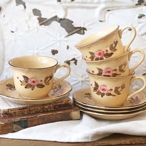 May include: A set of four vintage teacups and saucers with a floral pattern. The cups and saucers are a pale yellow color with a brown rim and a floral design featuring pink roses and brown leaves. The cups and saucers are stacked on top of each other on a white linen tablecloth.