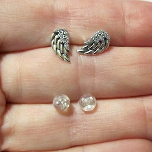 May include: A pair of silver-toned wing-shaped stud earrings with clear gemstone accents. The earrings are displayed with two clear earring backs. The wings have detailed feathering and are slightly curved.