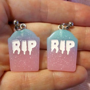 May include: A pair of pastel pink and blue tombstone earrings with the letters "RIP" in white. The earrings have a glittery finish and are attached to silver-colored earring hooks.