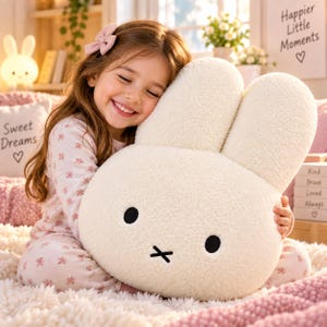 May include: A large, cream-colored plush pillow shaped like a rabbit head with two tall ears, two round black eyes, and a black cross-shaped mouth. The pillow is made of a soft, textured, fleece-like material and features a simple, minimalist design.