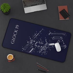 May include: A rectangular dark blue desk mat featuring a white technical schematic diagram of a Glock 19 firearm with numbered parts. The text GLOCK 19 is printed in white on the left side. The mat has a smooth surface and rounded corners.