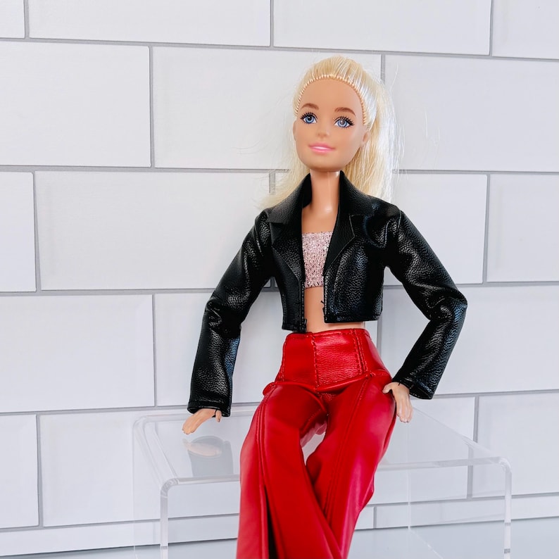 May include: A fashion doll with blonde hair and blue eyes, dressed in a black faux leather jacket, a pink sequined crop top, and red trousers. The doll is seated on a clear acrylic surface.
