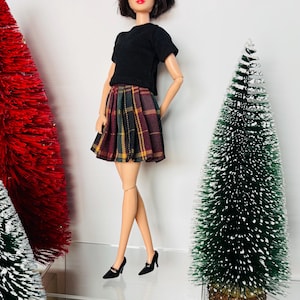 May include: A doll wearing a black short-sleeved top and a plaid skirt in shades of burgundy, green, and yellow. The doll is also wearing black heels. Two small Christmas trees are in the background.