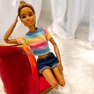 May include: A fashion doll with brown hair and skin, wearing a rainbow striped t-shirt and denim shorts. The doll is posed on a red chair, with one arm resting on its head. The doll is a popular toy.