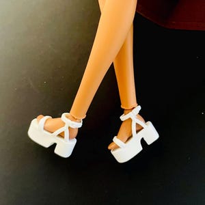 May include: White platform sandals with ankle straps. The sandals have a chunky sole and a minimalist design. The image shows the sandals on a doll's legs, with a dark background.