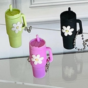 May include: Three miniature tumbler keychains in pastel colors: light green, lavender, and black. Each tumbler features a white daisy and a silver chain. The tumblers have a handle and a straw.