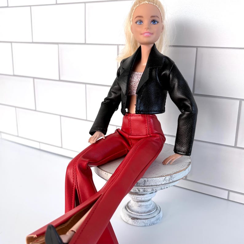 May include: A doll dressed in a black faux leather jacket, a sparkly crop top, and red split-leg trousers. The doll is seated on a small, white, distressed pedestal. The doll has blonde hair and is wearing black shoes.