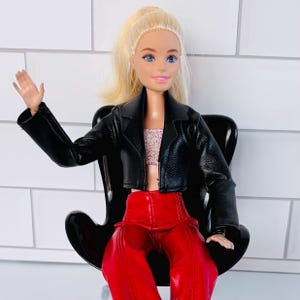 May include: A doll with blonde hair and blue eyes, dressed in a black leather jacket, a pink top, and red trousers. The doll is seated in a black chair with a curved back. The background is a white tiled wall.