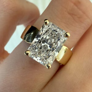 Op de afbeelding: Een ring met een heldere edelsteen in radiant-slijpvorm, gezet in een gepolijste geelgouden band. De rechthoekige steen wordt op de hoeken vastgehouden door vier gouden klauwen en heeft een briljant gefacetteerd oppervlak dat licht reflecteert.