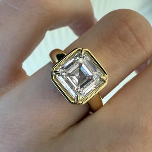 Puede incluir: Un anillo de oro con una gran piedra preciosa transparente de corte cuadrado montada en un engaste de bisel de cuatro puntas. La banda es de oro amarillo liso y pulido, con un diseño geométrico que resalta la piedra central facetada.
