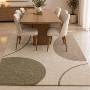 Könnte beinhalten: Ein rechteckiger Teppich mit cremefarbener Basis und großen, überlappenden kreisförmigen Mustern in gedämpftem Olivgrün und sanftem Taupe. Der Teppich hat einen strukturierten, flauschigen Flor und liegt unter einem Holzesstisch mit Stühlen.