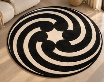 Op de afbeelding: Een rond vloerkleed met een contrastrijk zwart en crèmekleurig patroon. Het ontwerp bestaat uit vijf gebogen, komma-vormige zwarte segmenten die vanuit een centraal stervormig middelpunt naar buiten spiralen voor een hypnotiserend effect.