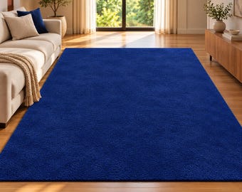 Op de afbeelding: Een groot, rechthoekig vloerkleed in een effen, diep koningsblauwe kleur met een dicht, pluizig en getextureerd oppervlak. Het kleed ligt plat op een houten vloer en bedekt een aanzienlijk deel van het zichtbare vloeroppervlak in de kamer.