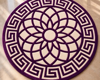 Op de afbeelding: Een rond vloerkleed in een dieppaarse en crèmekleurige kleurstelling. Het ontwerp bevat een centraal bloemachtig geometrisch patroon, omringd door een brede rand met een doorlopend Grieks sleutelmotief in paars en crème.
