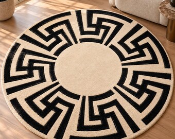 Op de afbeelding: Een rond vloerkleed met een crèmekleurige achtergrond en een opvallend, zwart geometrisch patroon. Het ontwerp bestaat uit een effen crèmekleurige middencirkel omringd door herhalende, in elkaar grijpende zwarte hoekige vormen als rand.