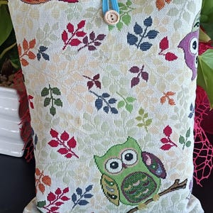 Peut inclure: Une pochette en tissu rectangulaire présentant un motif tissé de hiboux colorés et de feuilles d'automne sur un fond beige clair. La pochette est fermée par une boucle élastique bleu clair et un petit bouton en bois gravé Handmade.