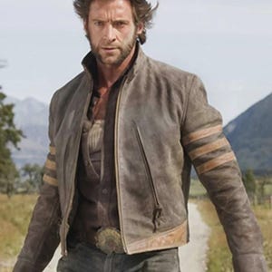 Könnte beinhalten: Eine braune Lederjacke mit Stehkragen, Frontreißverschluss und drei horizontalen beigen Streifen an jedem Ärmel. Die Jacke wird über einem dunkelbraunen Hemd mit Knopfleiste und einer dunklen Jeans mit einer großen, verzierten Metallschnalle getragen.