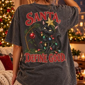 Op de afbeelding: Een antracietgrijs t-shirt met een afbeelding van een zwarte kat verstrikt in kleurrijke kerstverlichting, met een gouden ster op de kop en reikend naar een rode kerstbal. De tekst luidt SANTA, DEFINE GOOD.