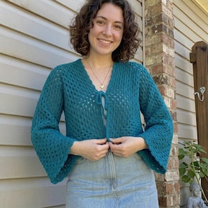 May include: A teal crochet cardigan featuring a V-neckline, long bell sleeves, and a front tie closure. The garment is crafted with an openwork stitch pattern, creating a textured, lightweight appearance. The cardigan is worn over a light-colored top.