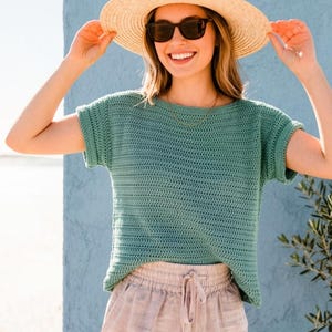 May include: A sage green short-sleeved crochet top with a round neckline and cuffed sleeves, paired with light beige drawstring shorts, a wide-brimmed straw hat, and dark-framed sunglasses, creating a coordinated ensemble for warm weather.
