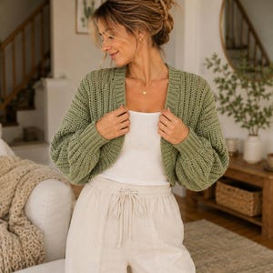 May include: A sage green open-front knit cardigan with long sleeves and ribbed cuffs, worn over a plain white tank top and light beige drawstring trousers. The cardigan features a textured, loose-knit pattern throughout the body and sleeves.