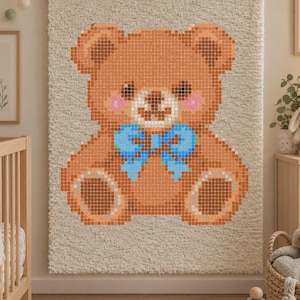 Op de afbeelding: Een rechthoekig wandkleed met een getextureerde crèmekleurige achtergrond en een gepixeld bruin teddybeerontwerp. De beer heeft donkerbruine oren, roze wangen en een felblauwe strik om de nek, alles weergegeven in een rasterpatroon.