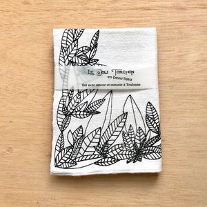 May include: White kitchen towel with a black and white leaf design. The towel has a label that reads "Le Joli Torchons en beau tissu fait avec amour et minutie à Toulouse".